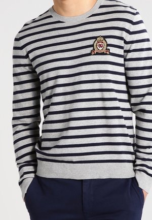 Mężczyzna w szarym i czarnym swetrze w poziome paski z małym haftowanym herbem na lewej piersi, w połączeniu z ciemnoniebieskimi spodniami.