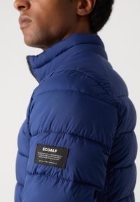 Dunkelblaues Steppjacke mit hohem Kragen, ausgestattet mit einem gesteppten Muster und einem schwarzen Ecoalf-Logoetikett am Ärmel. Leichtes Material.