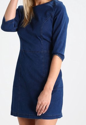 Femme portant une robe en denim bleu foncé ajustée avec des manches trois-quarts, debout devant un fond clair uni.