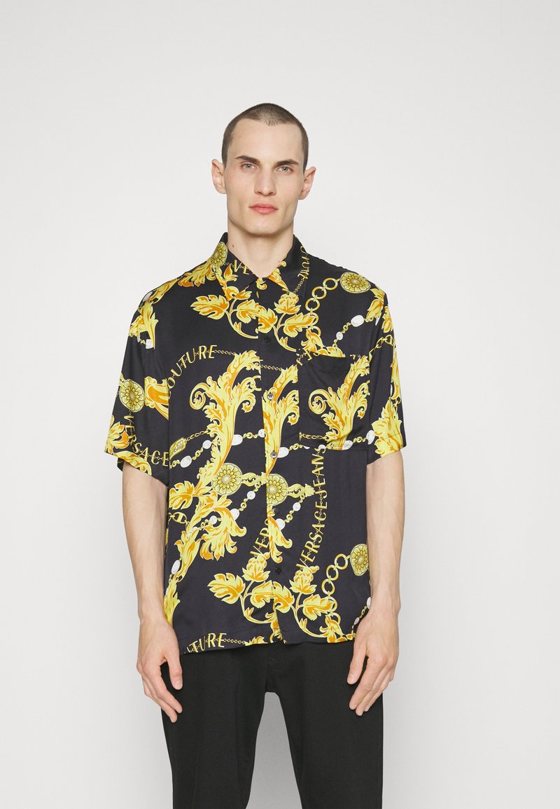 Chemise noire à manches courtes avec un motif baroque doré et du texte. Le tissu semble léger, avec une coupe décontractée et un col boutonné.