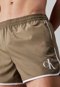 Pantaloni da bagno marroni con vita elasticizzata, accenti laterali bianchi e un logo "CK" bianco nella parte inferiore destra. Texture liscia.