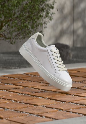 Λευκό αθλητικό παπούτσι Tommy Hilfiger με χοντρή σόλα και κορδόνια, τοποθετημένο πάνω σε ξύλινο δάπεδο με πετρούλες και σχέδια σε υπαίθριο χώρο.