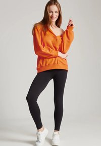 Pomarańczowy sweter z dekoltem w serek, bufiastymi rękawami i prążkowanym dołem, połączony z czarnymi legginsami i białymi sneakersami.