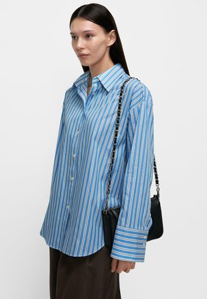 Kobieta w za dużej, niebiesko-białej koszuli w paski z długimi rękawami, niosąca małą czarną torebkę na łańcuszku.