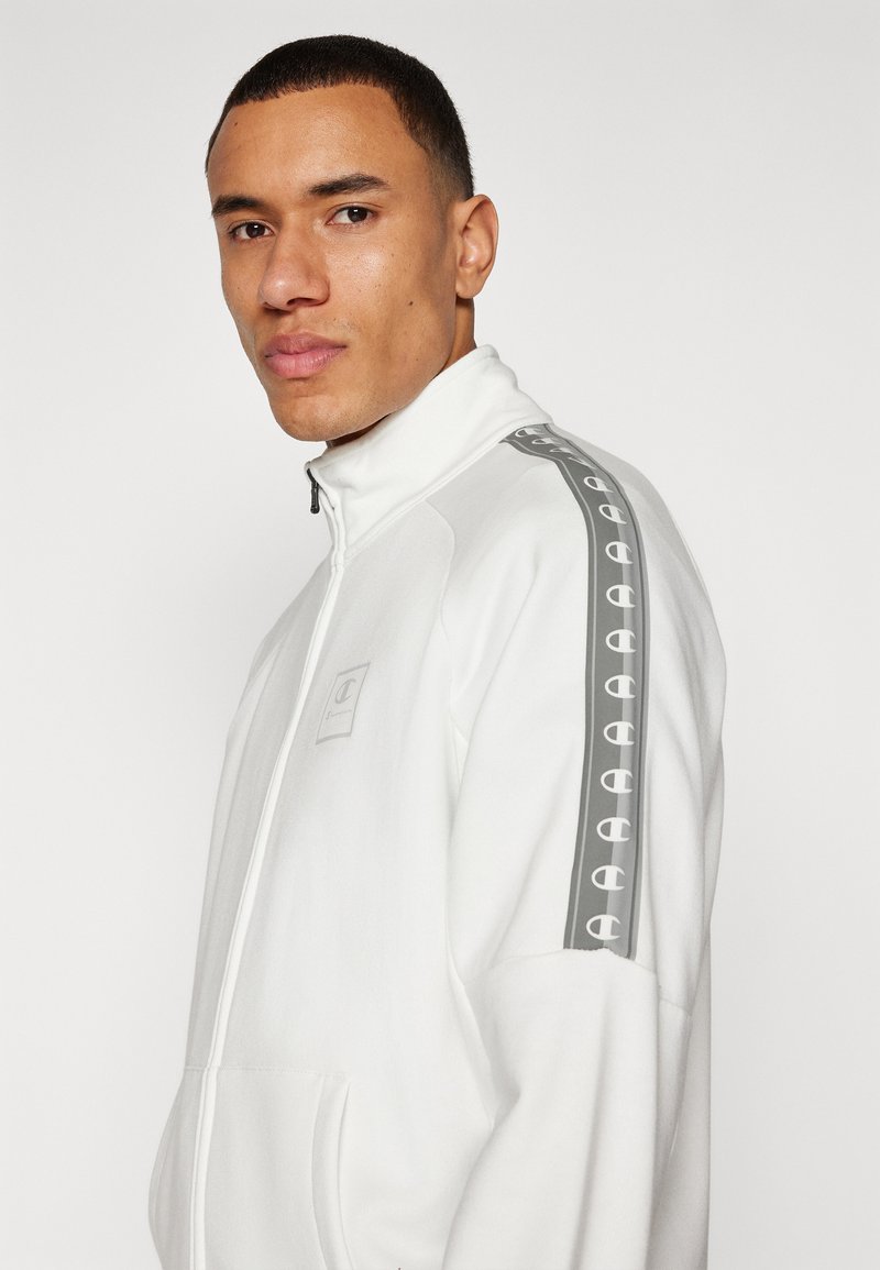 Giacca bianca con zip e collo alto, caratterizzata da strisce laterali grigie con accenti del logo e tasca frontale sul lato sinistro. Texture liscia.