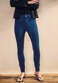 Tmavomodré skinny džínsy s vysokým pásom, vyrobené z elastického denimu, s predným zapínaním na gombík a jemným vyblednutím. Kombinujte s čiernymi šľapkami.