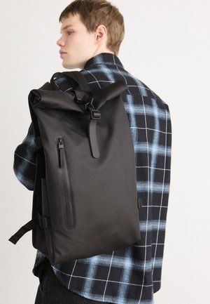 Black roll-top backpack made of waterproof material, featuring a side zipper pocket and adjustable straps, worn over a blue plaid shirt.