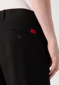 Pantaloni negri cu închidere prin nasture, buzunare laterale și un buzunar posterior cu un mic etichetă roșie "HUGO". Textură de țesătură netedă.