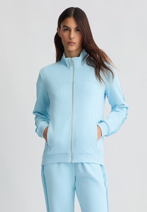 Felpa zip-up azzurra chiara con colletto alto, texture liscia e tasche laterali. Presenta inserti in rete lungo le maniche e pantaloni coordinati.