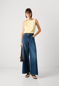 Top sem mangas amarelo claro com um detalhe de nó, combinado com jeans de denim azul de perna larga e cintura alta. Bolsa castanha e sapatos de salto alto.