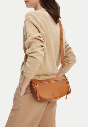 Mujer con suéter y pantalones beige, llevando un bolso cruzado de cuero marrón claro con cremalleras plateadas y la marca "JENNY".
