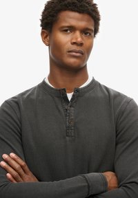 Mężczyzna ze złożonymi ramionami, ubrany w ciemnoszarą koszulę henley z długim rękawem z zapięciami na guziki, patrzący lekko w bok na białym tle.