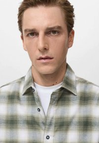 Camisa de cuadros en verde y blanco con cuello; presenta botones negros y una textura suave; usada sobre una camiseta blanca.