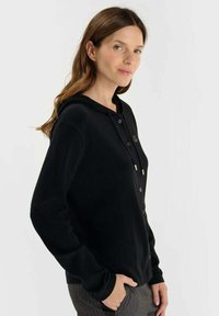 Zwarte hoodie cardigan gemaakt van zacht materiaal, met een knoopsluiting aan de voorkant en trekkoordaccenten. Voorzien van een ontspannen pasvorm.