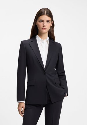 Femme aux longs cheveux bruns portant une veste et un pantalon de costume noirs ajustés sur une chemise blanche à col, sur fond blanc.