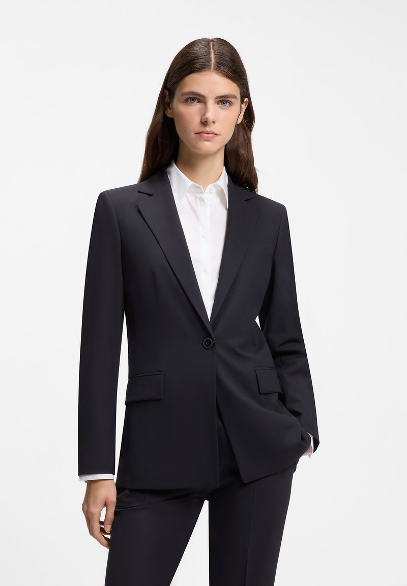Femme aux longs cheveux bruns portant une veste et un pantalon de costume noirs ajustés sur une chemise blanche à col, sur fond blanc.