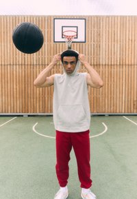 Grå ærmeløs hættetrøje med kængurulomme, røde sweatpants og hvide sneakers. En basketball i luften, træ-gymnastikbaggrund.