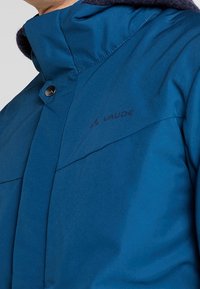 Gros plan sur une veste Vaude bleue avec un col montant et un bouton visible près de l'encolure.