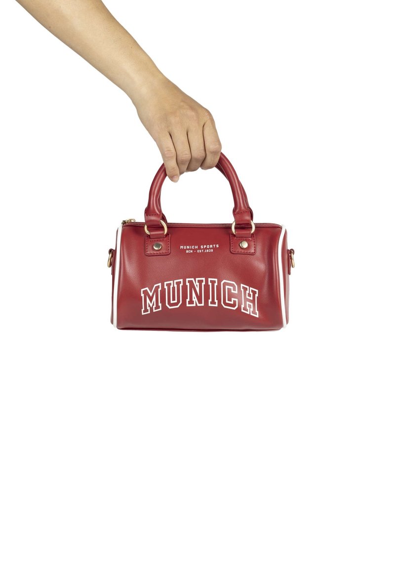 Main tenant un petit sac à main rouge avec double poignée, texte blanc "MUNICH" et "Munich Sports BCN - Est. 1939" à l'avant.