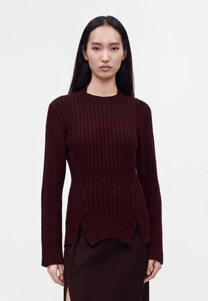 Femme aux longs cheveux noirs portant un pull côtelé bordeaux foncé et une jupe assortie sur un fond clair uni.