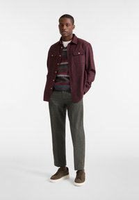 Maroon knoopjasje over een geblokte trui, gecombineerd met grijs geruite broek en bruine sneakers. Soepele texturen en casual ontwerp.