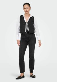 Zwarte cropped vest met strikdetail, geborduurd ontwerp en franjes over een wit overhemd, gecombineerd met zwarte straight-leg jeans en flats.