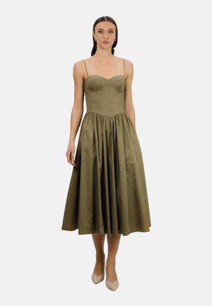 Kvinne iført olivengrønn ermeløs midikjole med tettsittende liv, fullt rynket skjørt og beige spisse hæler, stående mot hvit bakgrunn.