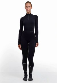 Justaucorps de sport noir sans coutures, manches longues, col zippé et coupe ajustée. Comprend des coutures blanches sur les jambes et une texture lisse.