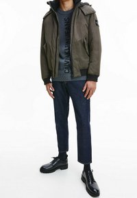 Zielona kurtka bomberka z kapturem, szary sweter z wyraźnym nadrukiem, niebieskie jeansy oraz czarne, sznurowane buty. Gładkie tekstury, swobodny styl.