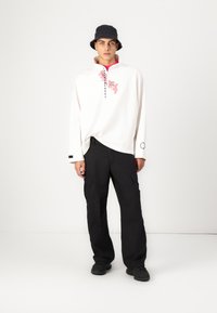 White pullover with a pink floral print and black text, paired with black cargo pants and a black bucket hat. Black sneakers complete the look.