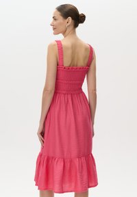 Różowa sukienka z marszczonym stanikiem i szerokimi ramiączkami. Posiada rozkloszowaną spódnicę z falbaną na dole oraz teksturowaną tkaninę. Pokazany widok z tylu.