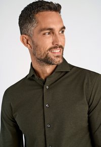 Man met kort donker haar en baard, gekleed in een donkergroen overhemd met knopen, lachend en kijkend naar de zijkant tegen een effen achtergrond.