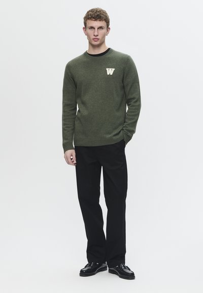 Pull en tricot vert avec un col rond et des poignets côtelés, arborant un logo "W" blanc. Associé à un pantalon noir et des chaussures noires.