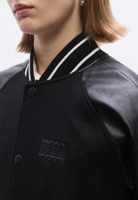 Jachetă varsity neagră cu mâneci din piele, guler striat cu dungulițe albe și o insignă cu logo pe piept. Textură netedă.