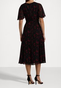 Svart midiklänning med röd blommönster, korta fladdrande ärmar, figursydd midja och en utsvängd kjol. Modellen bär svarta klackskor med remmar.