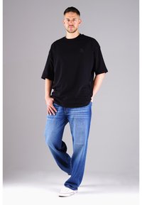Mann steht mit Händen in den Taschen, trägt ein schwarzes übergroßes T-Shirt, blaue Jeans und weiße Turnschuhe vor einem grauen Hintergrund.