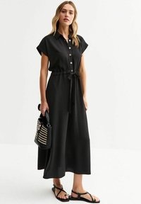 Vestido negro de botones con mangas cortas enrolladas, cintura entallada y un corte holgado; combinado con sandalias negras y un bolso a rayas.