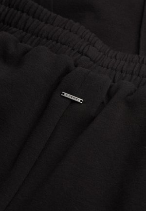 Zwarte katoenen joggers met een elastische tailleband en een klein metalen label met de tekst "Superdry" aan de voorkant. De textuur oogt zacht en glad.