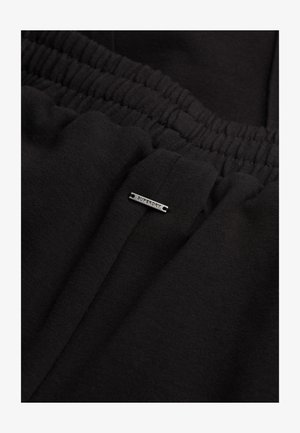 Zwarte katoenen joggers met een elastische tailleband en een klein metalen label met de tekst "Superdry" aan de voorkant. De textuur oogt zacht en glad.