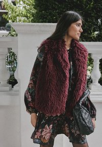 Weinroste Kunstpelz-Weste mit hohem Kragen, kombiniert mit einem dunklen Blumenkleid und einer strukturierten schwarzen Handtasche, vor weißem Hintergrund.