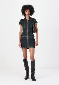 Denim kjole med glidelås foran, korte ermer og en figursydd silhuett. Har olivengrønne detaljer og en lomme. Bæres med høye svarte støvler.