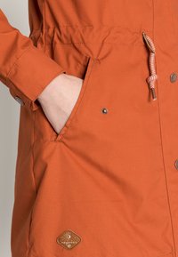 Pomarańczowa kurtka z troczkiem w talii, z zatrzaskami i metką. Posiada gładką teksturę i boczną kieszeń.