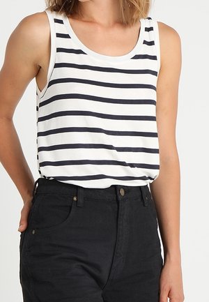 Osoba ubrana w bezrękawnikową białą bluzkę z czarnymi poziomymi paskami, wsuniętą w wysokowadze czarne spodnie.