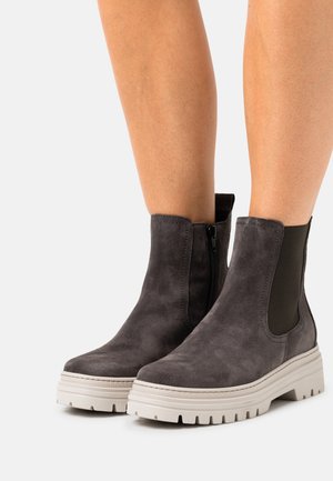 Bottines en suède gris avec une épaisse semelle blanche, fermeture éclair sur le côté et panneaux élastiques pour un enfilage facile. Surface texturée avec des lignes épurées et un design moderne.