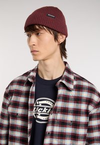 Tmavě červená pletená čepice má ribbový povrch a černou nášivku. Nosit se s kostkovanou flanelovou košilí v červených, modrých a béžových tónech.
