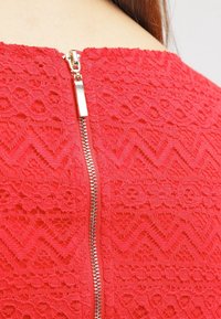 Vêtement en dentelle rouge avec une fermeture éclair en métal argenté visible dans le dos. La texture comprend des motifs en zigzag et floraux.