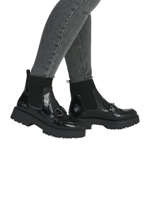 Jambes vêtues d'un jean skinny gris et de bottines noires brillantes à semelles épaisses et poignets en tricot texturé, debout les pieds croisés.