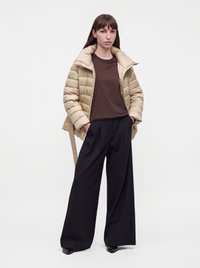 Frau trägt eine beige Steppjacke über einem braunen Hemd, schwarze weit geschnittene Hosen und schwarze spitze Schuhe, steht mit den Händen in den Taschen.