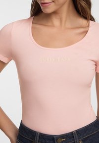 Top rosa a maniche corte con scollo rotondo e una texture liscia, con il logo "GUESS JEANS" in oro chiaro sul petto.