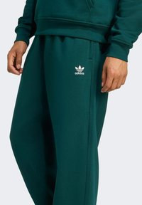 Groene Adidas-sweatpants van zacht materiaal, met een elastische tailleband, zijzakken en een wit logo op de linkerbovenheup.